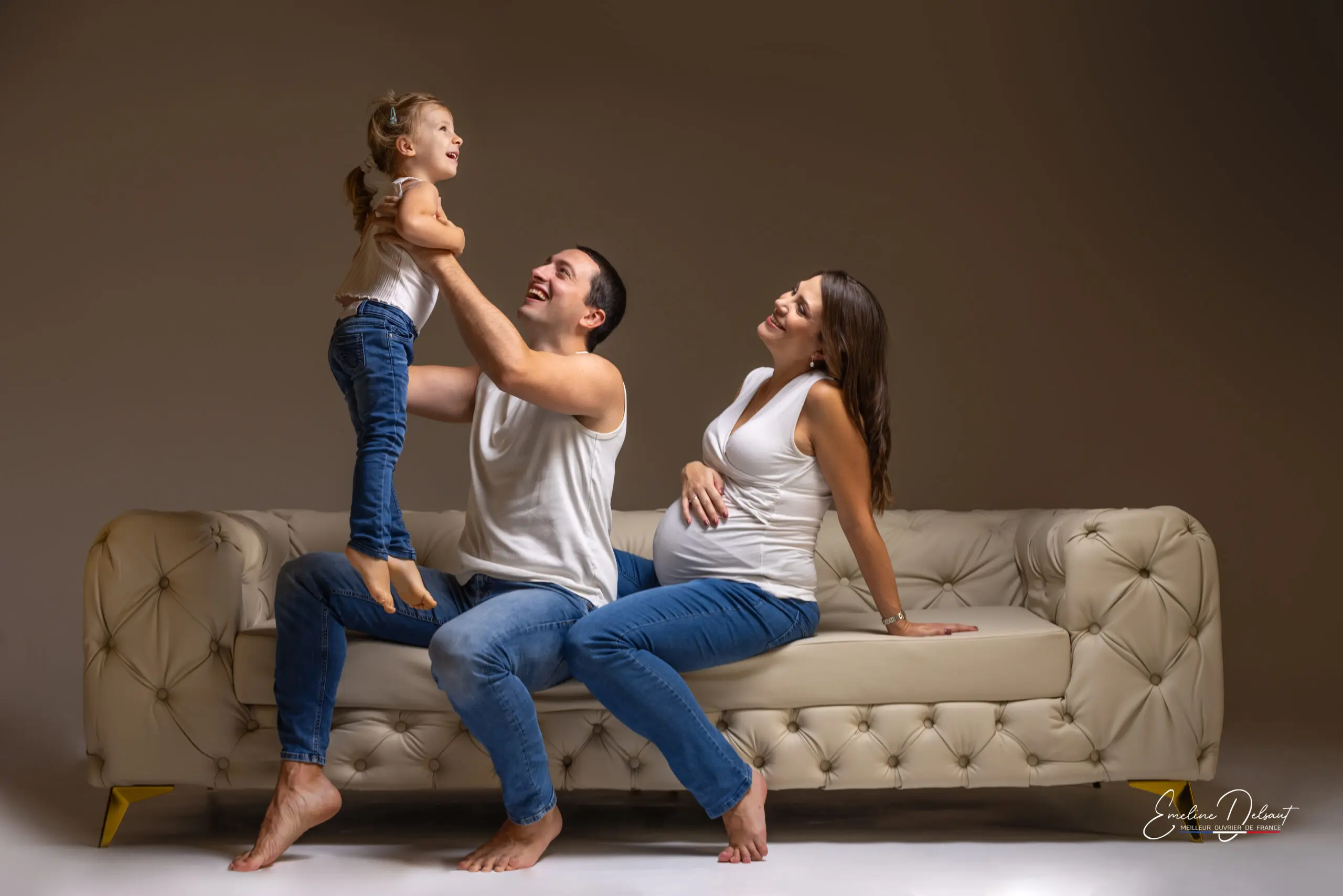 Photo de famille dans un superbe canapé au Studio END. Le papa et la fille jouent et maman souris avec une main sur le ventre ou grandi son bébé